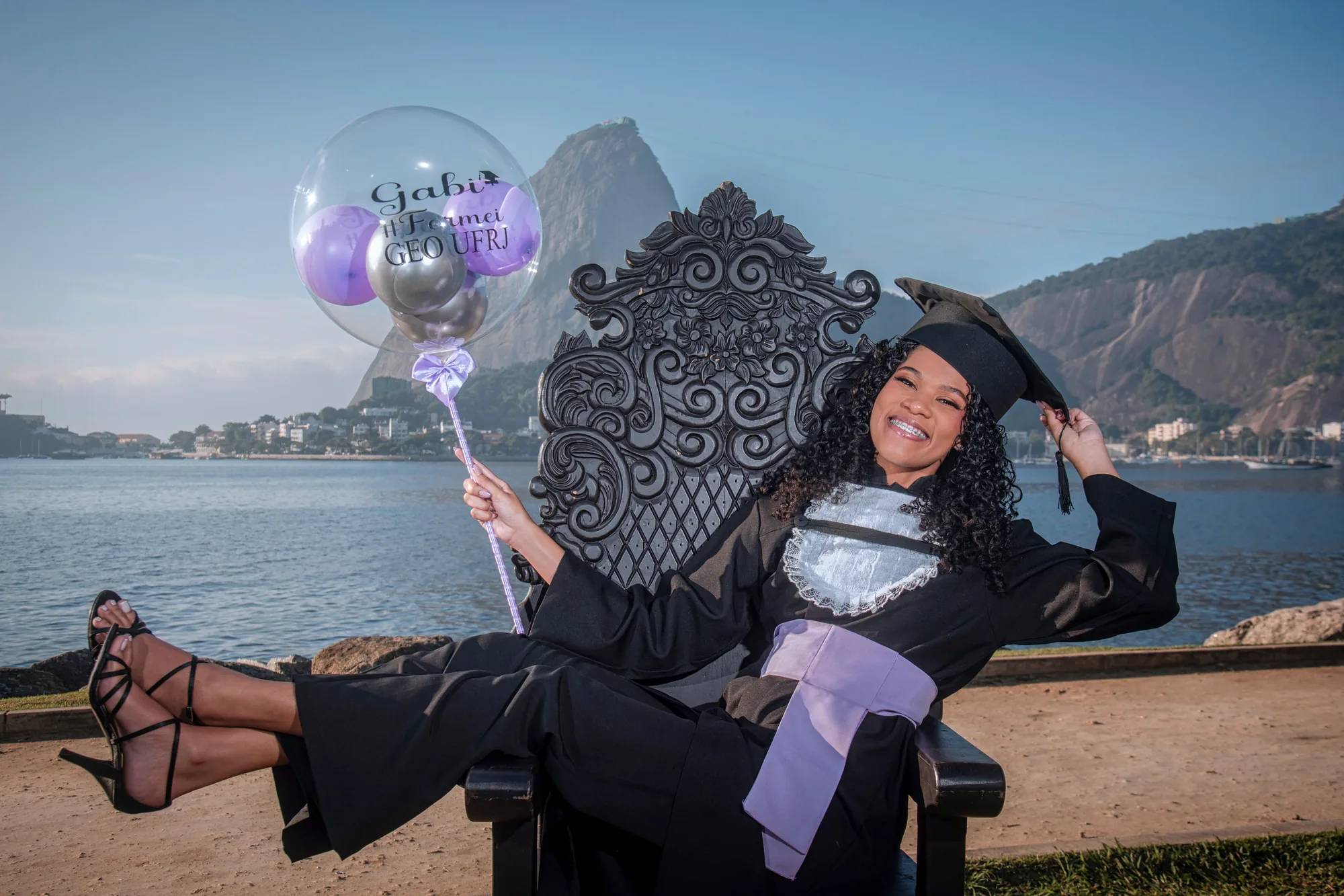 Formanda de beca na cadeira entalhada com um balão personalizado, diante do Pão de Açúcar
