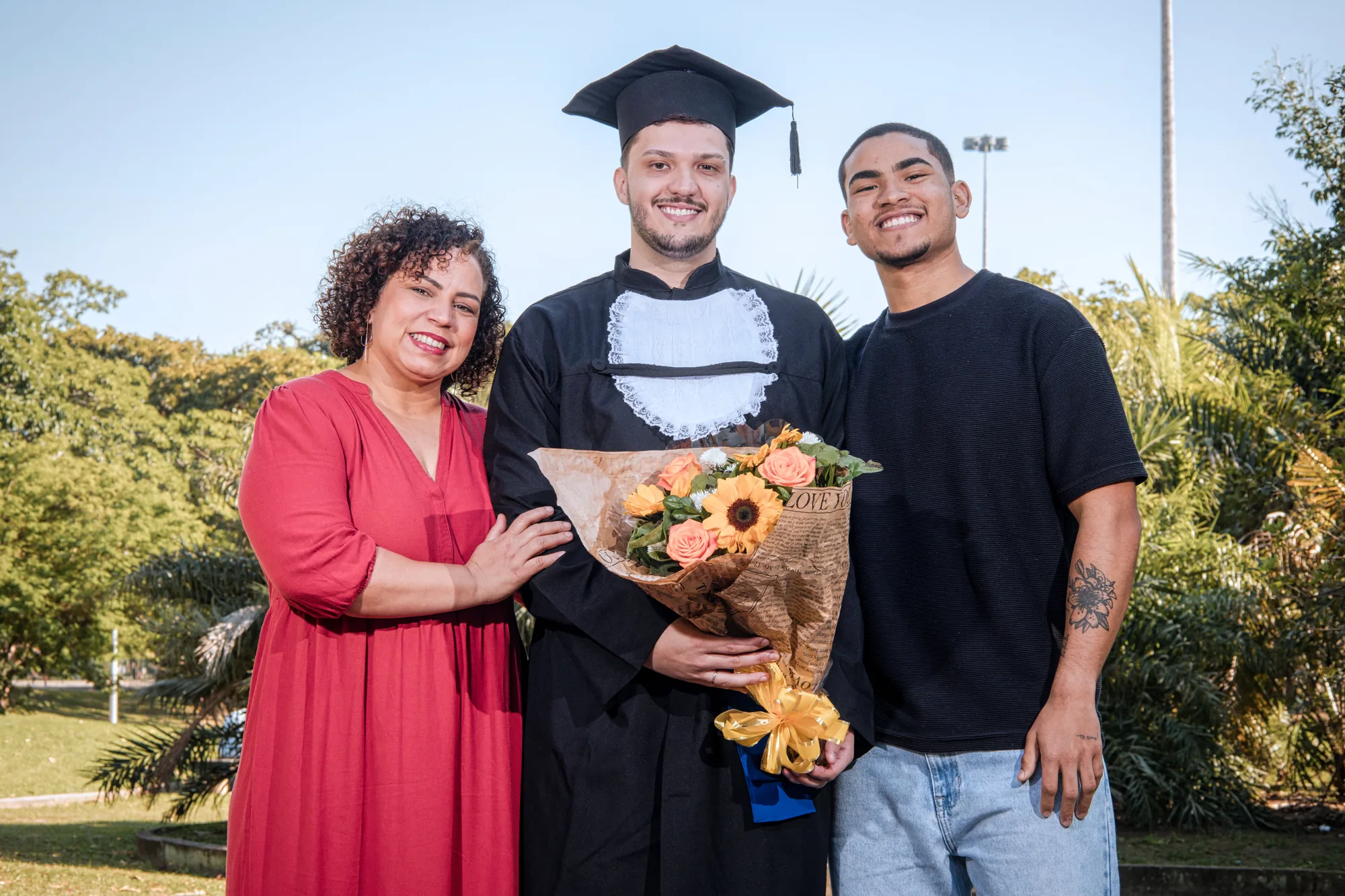 Formando de beca com a mãe e um amigo, segurando um buquê de flores no Aterro do Flamengo