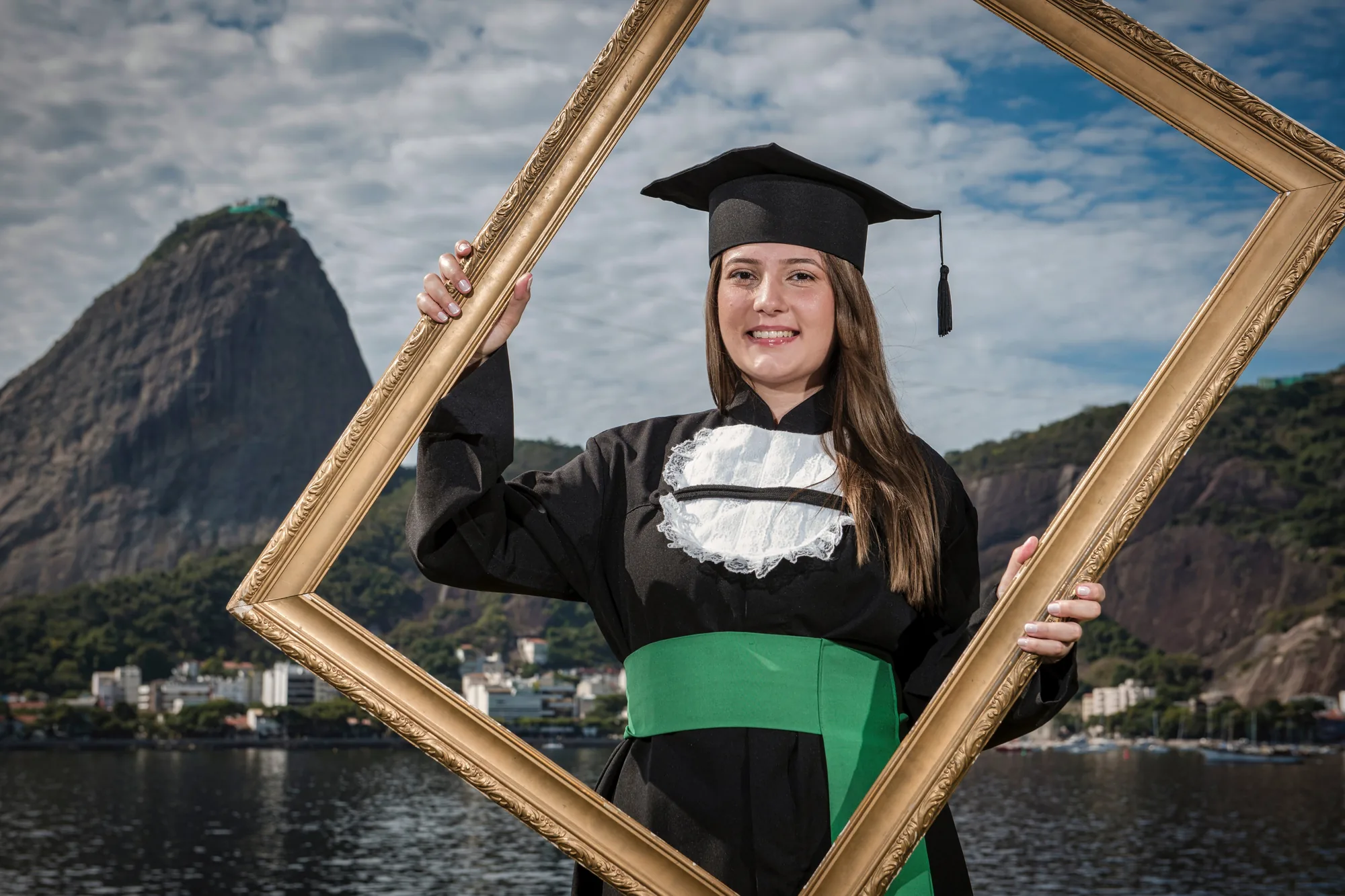 Formanda de beca sorri dentro de uma moldura dourada, com o Pão de Açúcar ao fundo