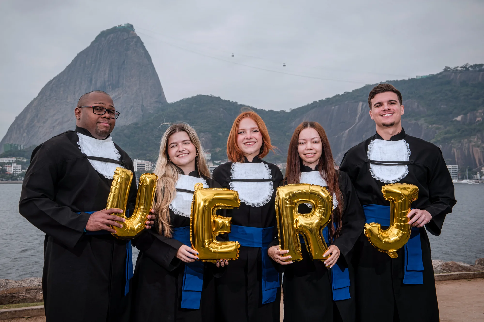 Grupo de formandos de beca segura balões dourados com as letras UERJ diante do Pão de Açúcar