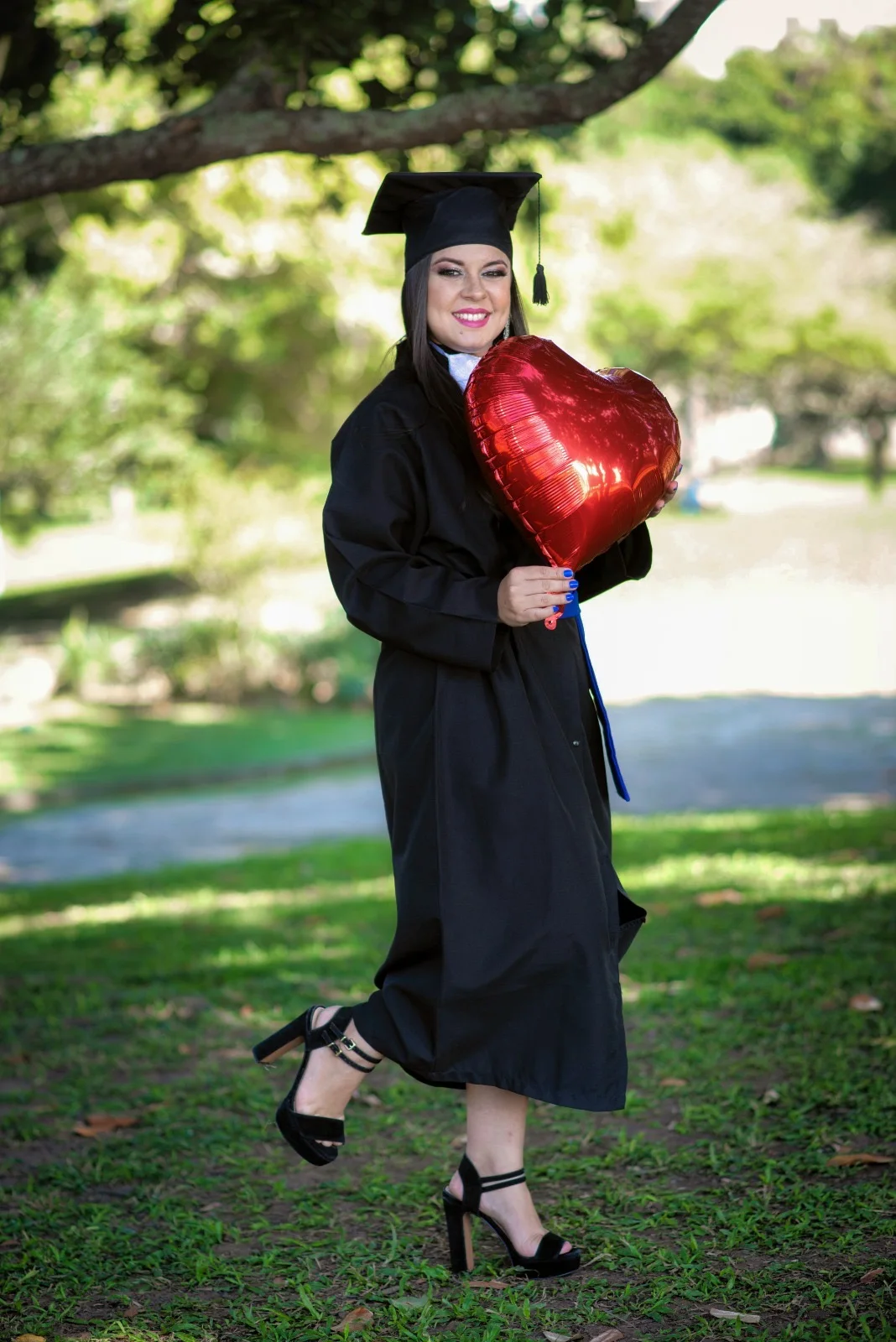 Formanda de beca posa com um balão de coração vermelho sob as árvores