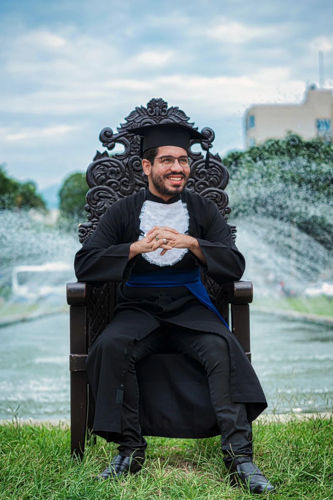 Formando de beca sorri sentado na cadeira entalhada diante de um chafariz