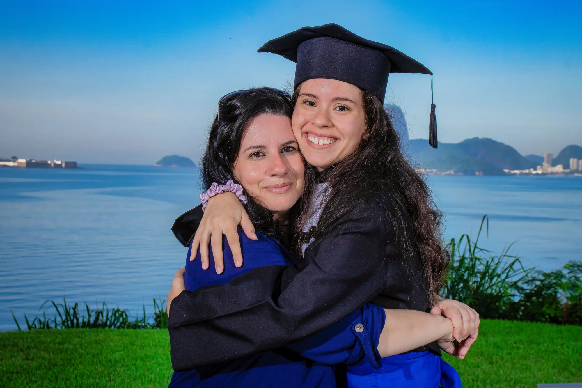 Formanda de beca abraça a amiga no gramado do MAC, com a Baía de Guanabara ao fundo