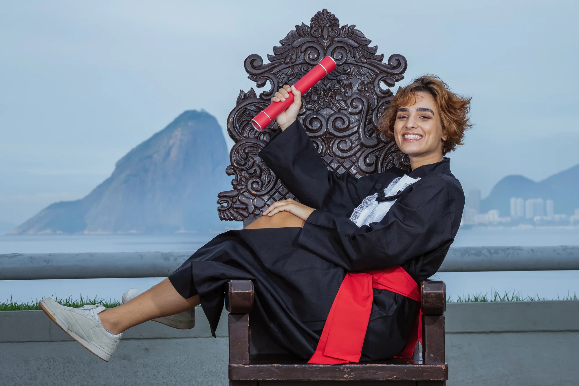 Formanda de beca ergue o canudo na cadeira entalhada, com o Pão de Açúcar ao fundo, no MAC de Niterói