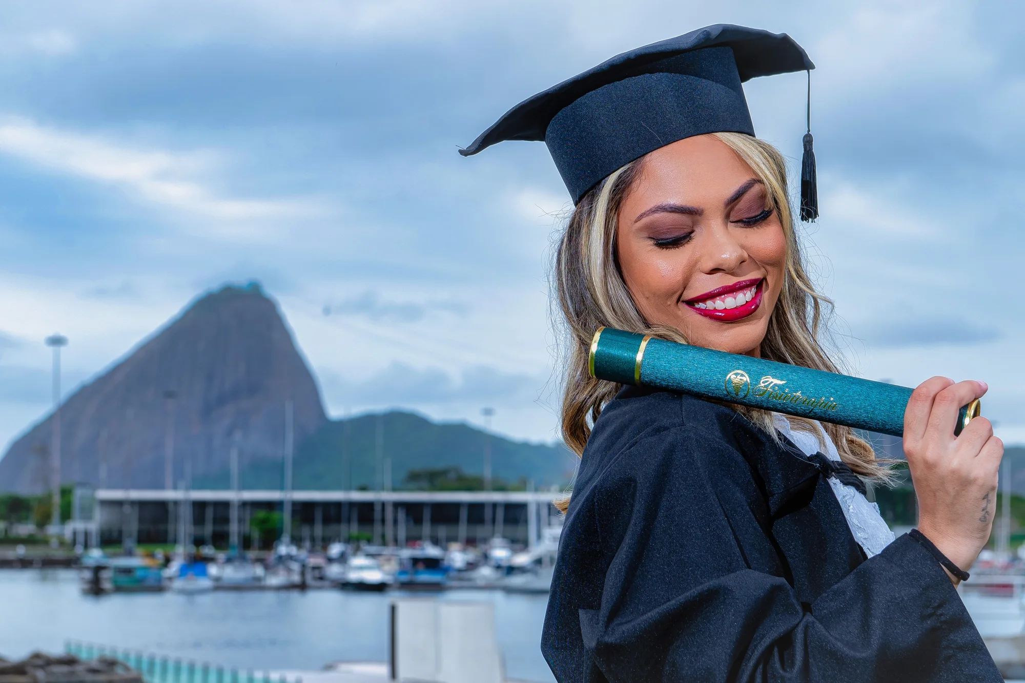 Formanda de beca sorri com o canudo, com a Marina da Glória e o Pão de Açúcar ao fundo