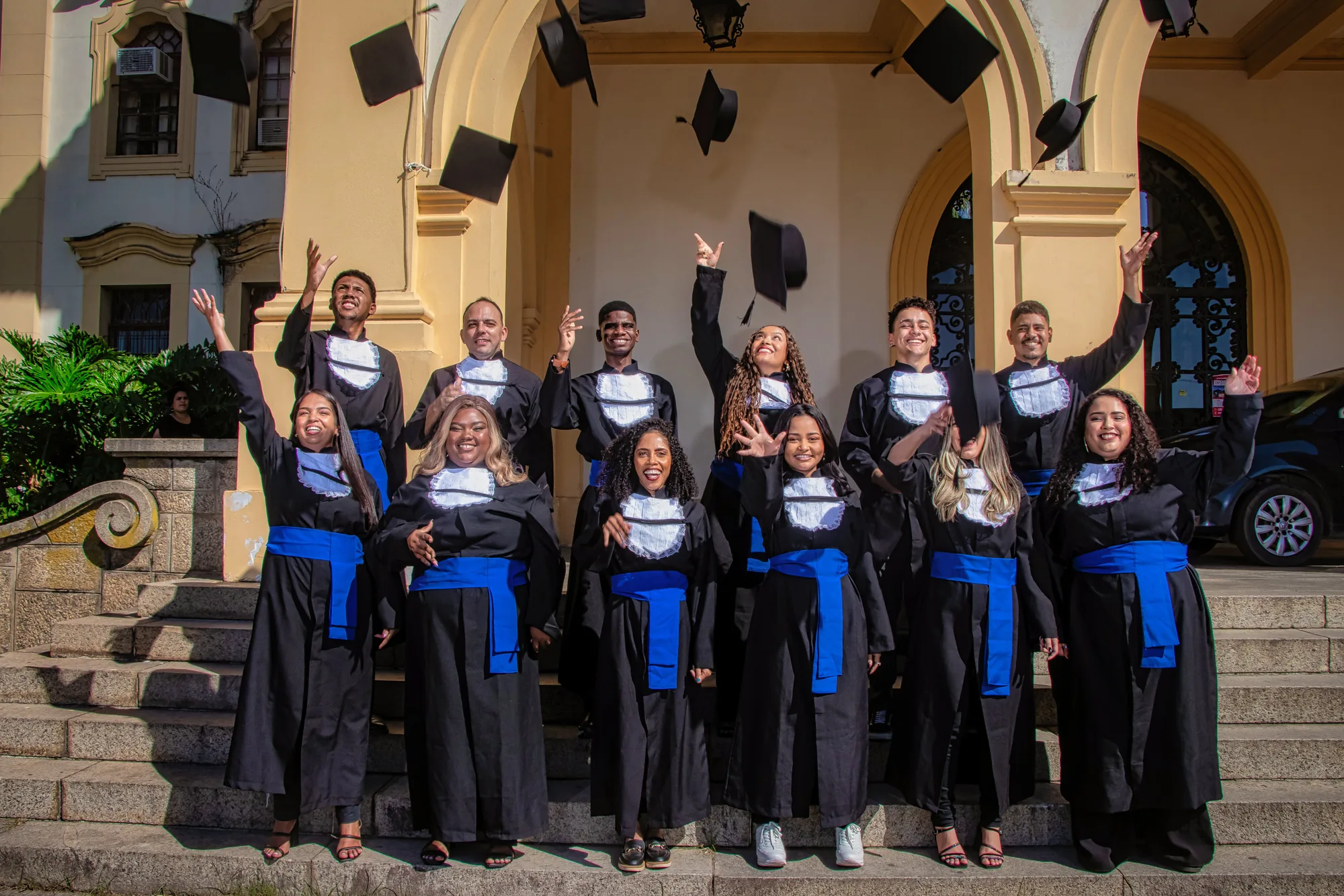 Turma de formandos joga os capelos para o alto na escadaria de um prédio histórico da UFRRJ