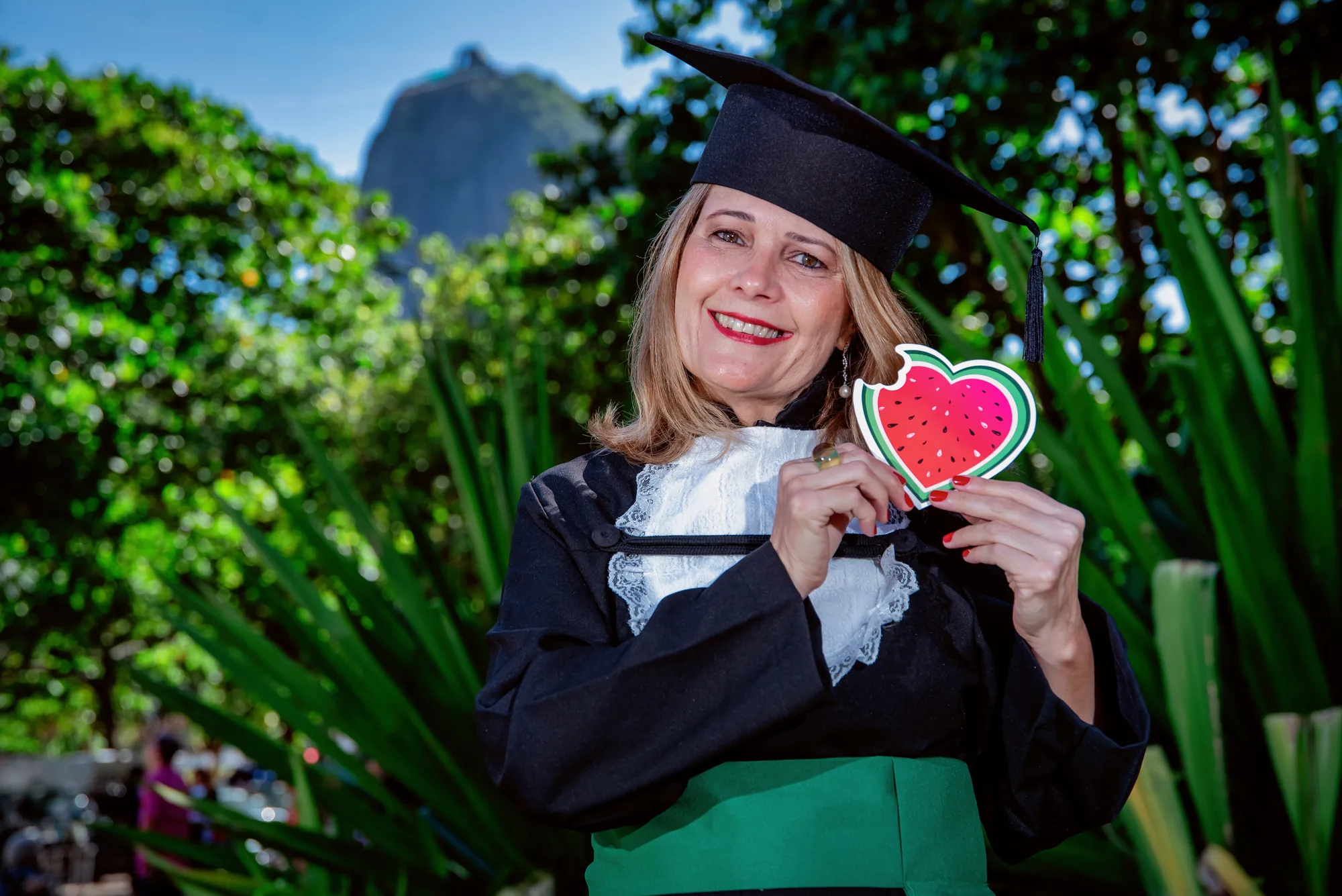 Formanda de beca e capelo sorri segurando um coração de melancia entre as plantas, na Urca