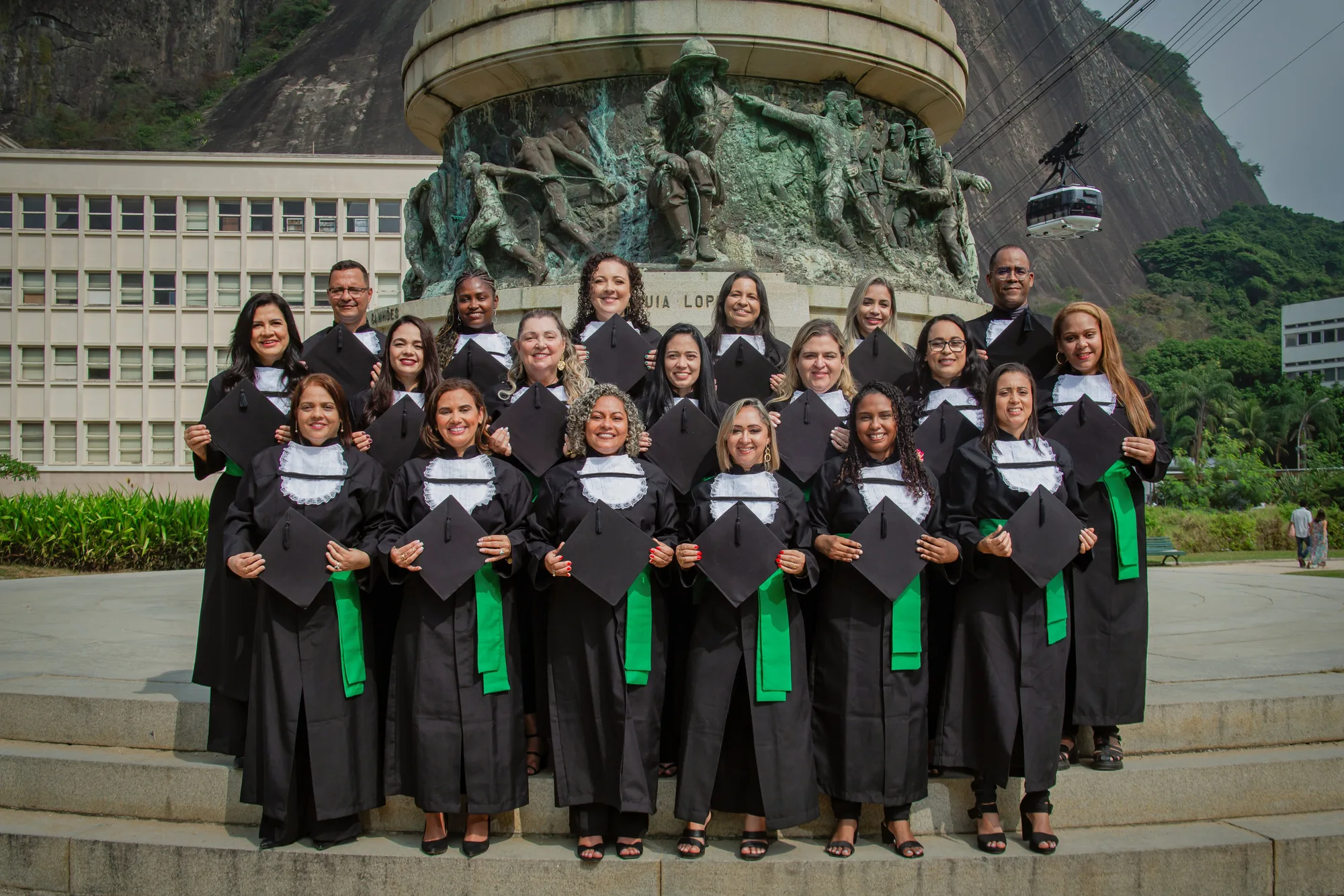 Turma de formandos de beca reunida diante do monumento, na Urca
