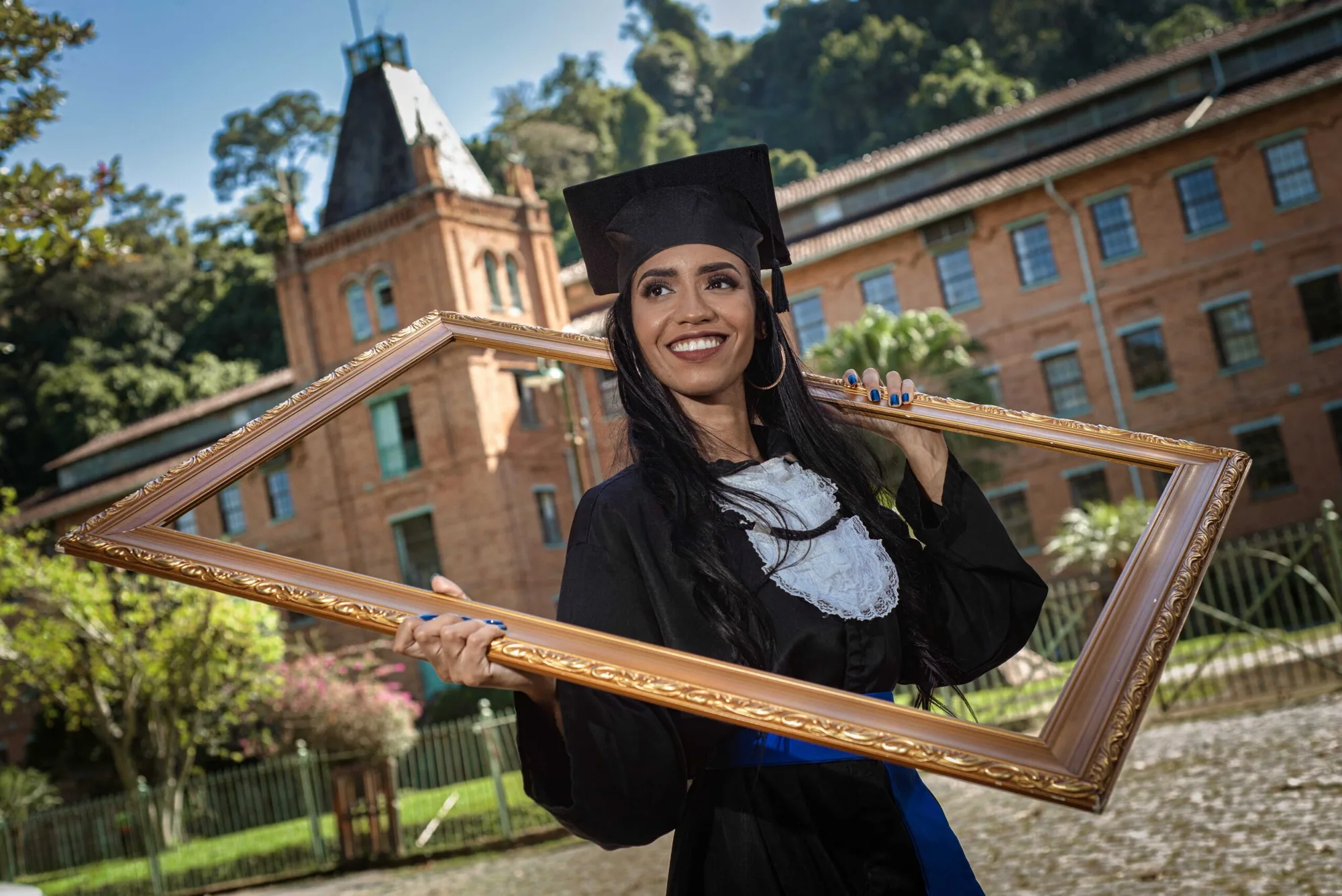 Formanda de beca segura uma moldura dourada diante de um prédio histórico