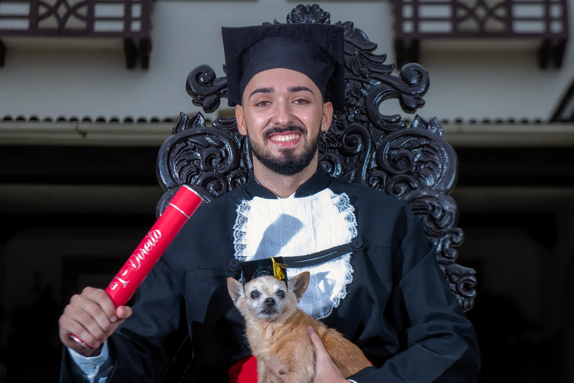 Formando de beca sorri na cadeira colonial segurando o canudo e o seu cachorro